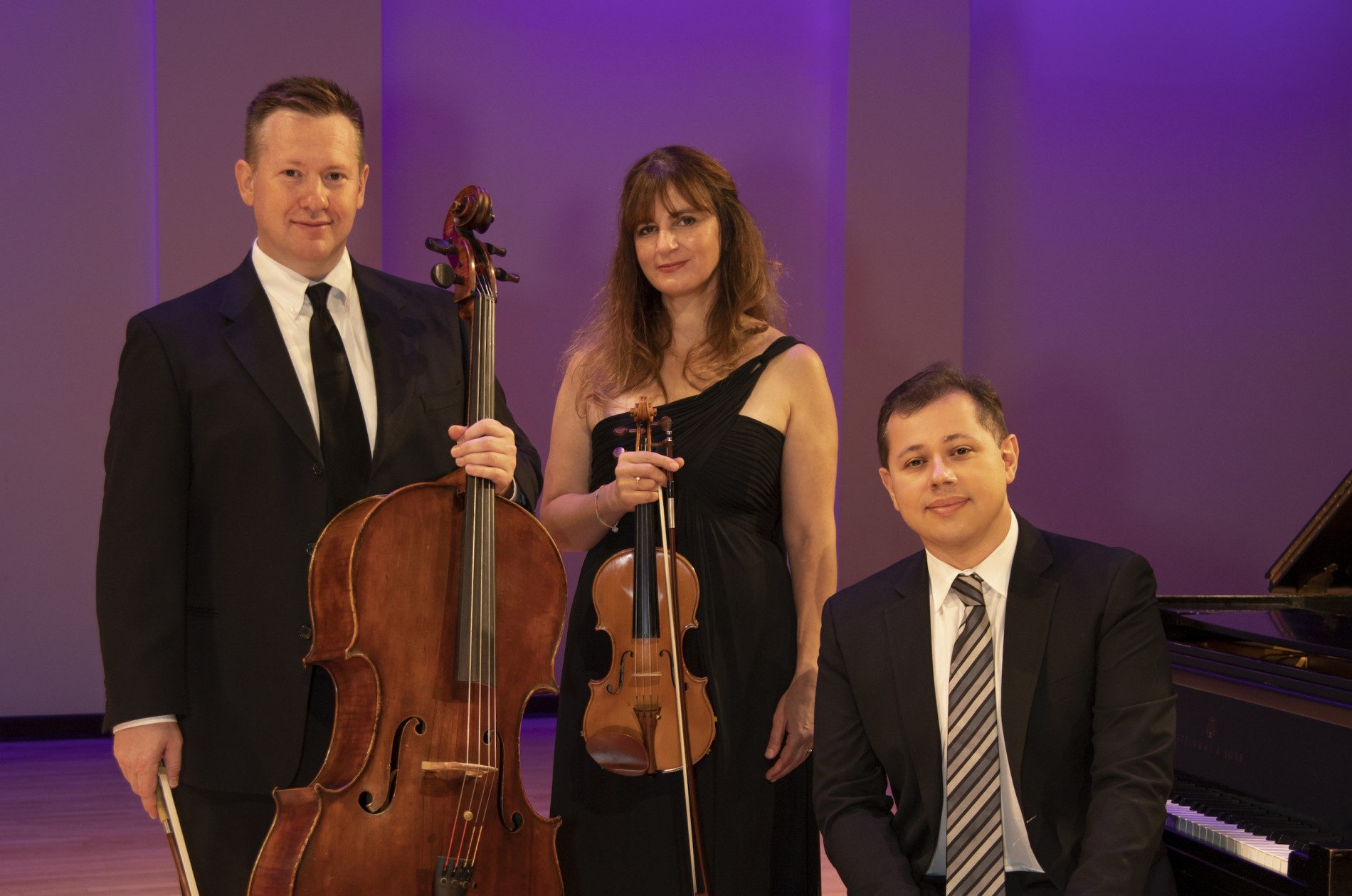 A formal portrait of a standing cellist, violinist, and pianist next to a piano.