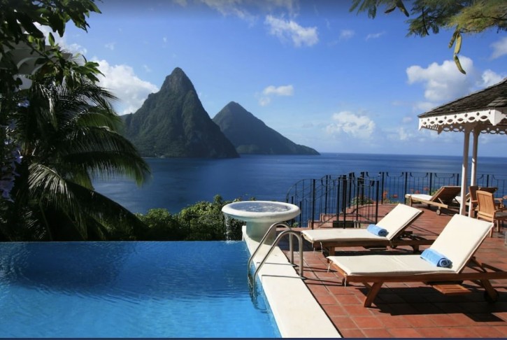 A poolside view of the St. Lucia vacation home. To the right are two lounge chairs. In the background are mountain peaks and the ocean.