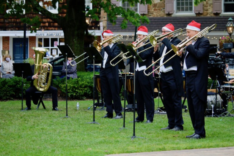 4 brass players stand and play their instruments on grass, wearing Santa Hats. There is another brass player to their left who is seated.