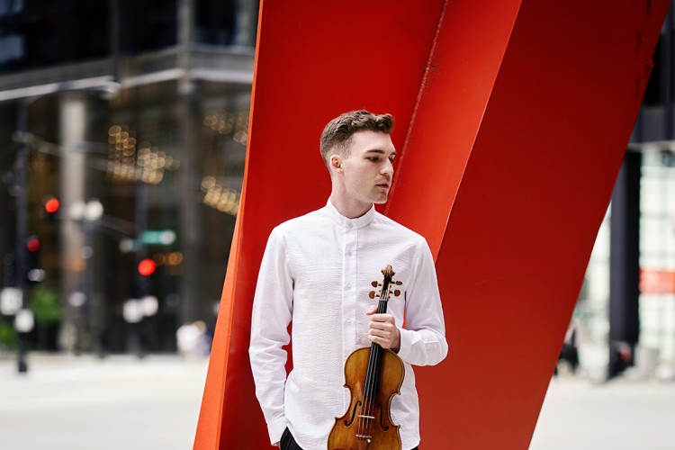 Alexi Kenney stands in front of a red sculpture. Alex is wearing a white long-sleeved shirt and black pants. One hand is in his pocket and the other is holding a violin.