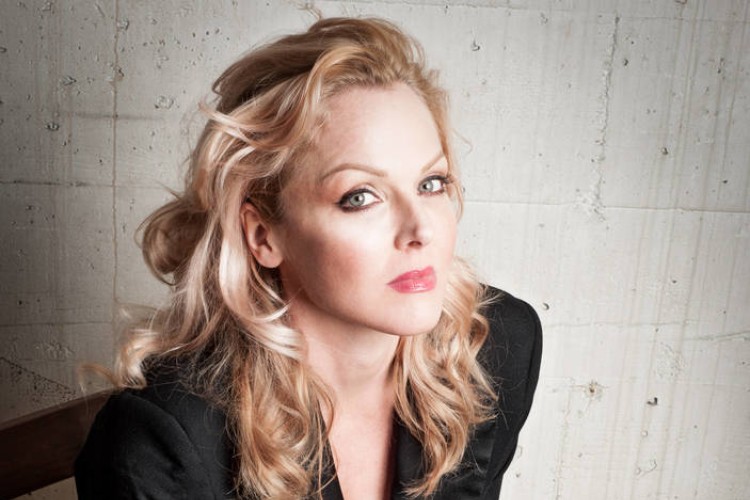 Singer Storm Large, seated on a chair in front of a concrete wall, looking at the camera. She is wearing a black blazer and black pants.