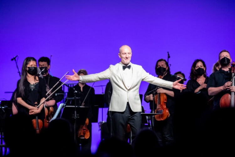 Music Director Rossen Milanov stands on stage facing the audience, arms spread wide. Behind him are the members of the orchestra, also standing and facing the audience.