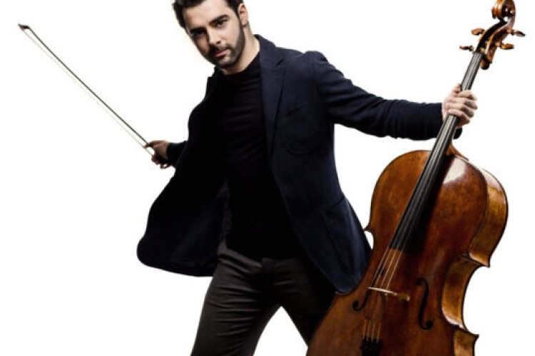 Cellist Pablo Ferrandez stands facing the camera. He is holding the neck of a cello in his left hand, and the bow in the right.