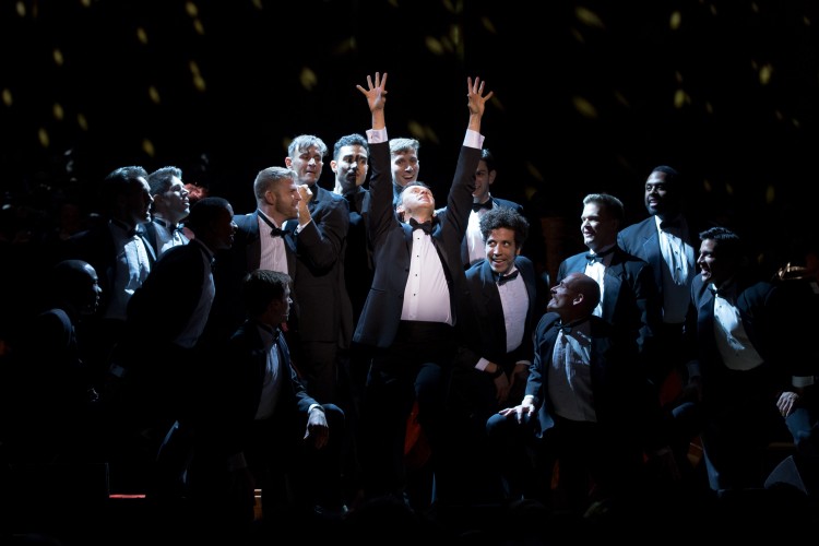 Men in tuxedos are clustered around the center of a stage, looking at one man in the middle. This man has his arms stretched above his head and is looking up.