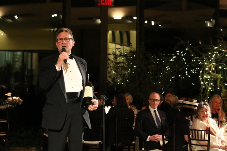A photo from a previous gala. The auctioneer is holding a microphone in his right hand and a bottle of wine in his left. Gala attendees are visible behind him.