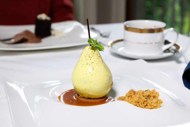 A pale green-yellow pear topped with a mint garnish sits on a pool of caramel next to a mound of brown sugar; a cup and saucer and other dessert lay in the background.