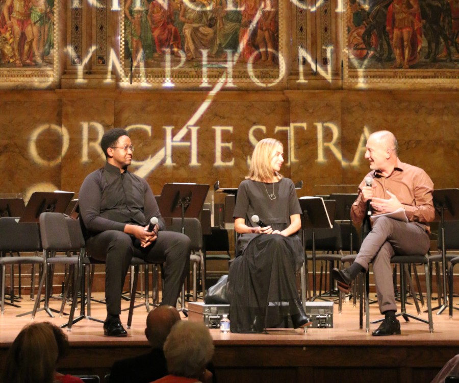 Three people sitting on a stage holding microphones, with projected text on the wall behind: Princeton Symphony Orchestra