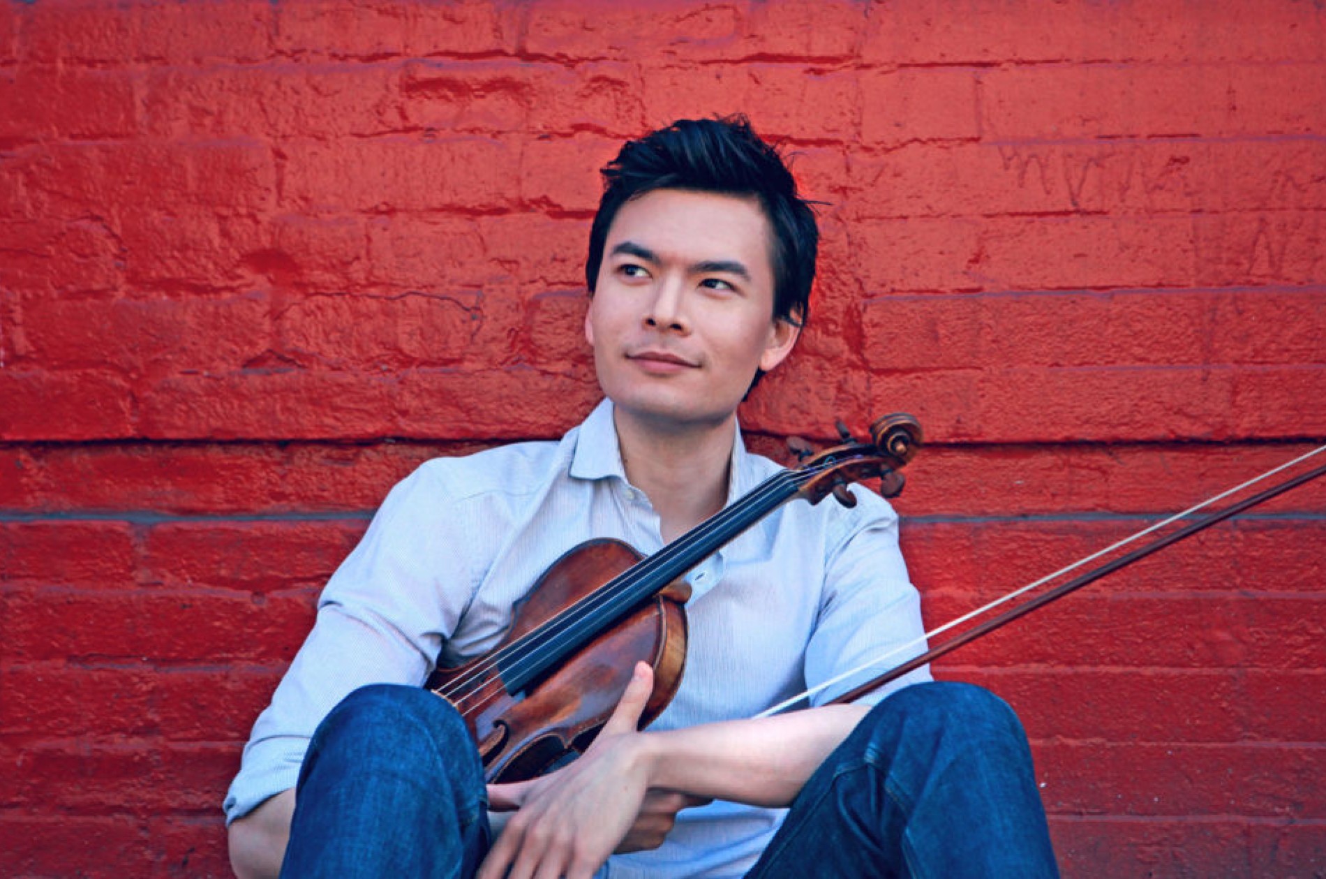 Stefan Jackiw holding a violin while sitting in front of a painted red brick wall