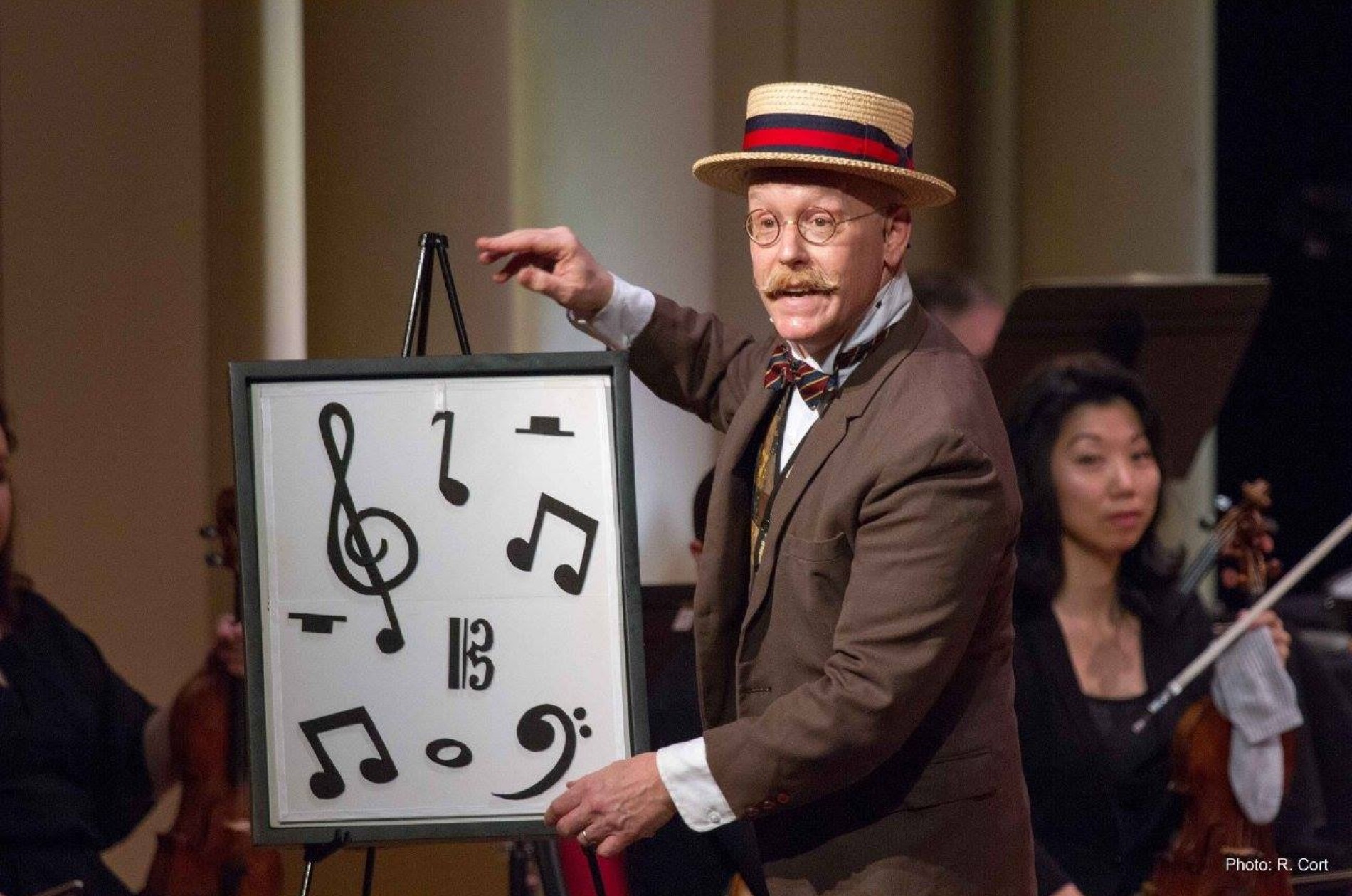Michael Boudewyns wears and brown suit and boater hat as he points out musical notes on an easel display in front of string players