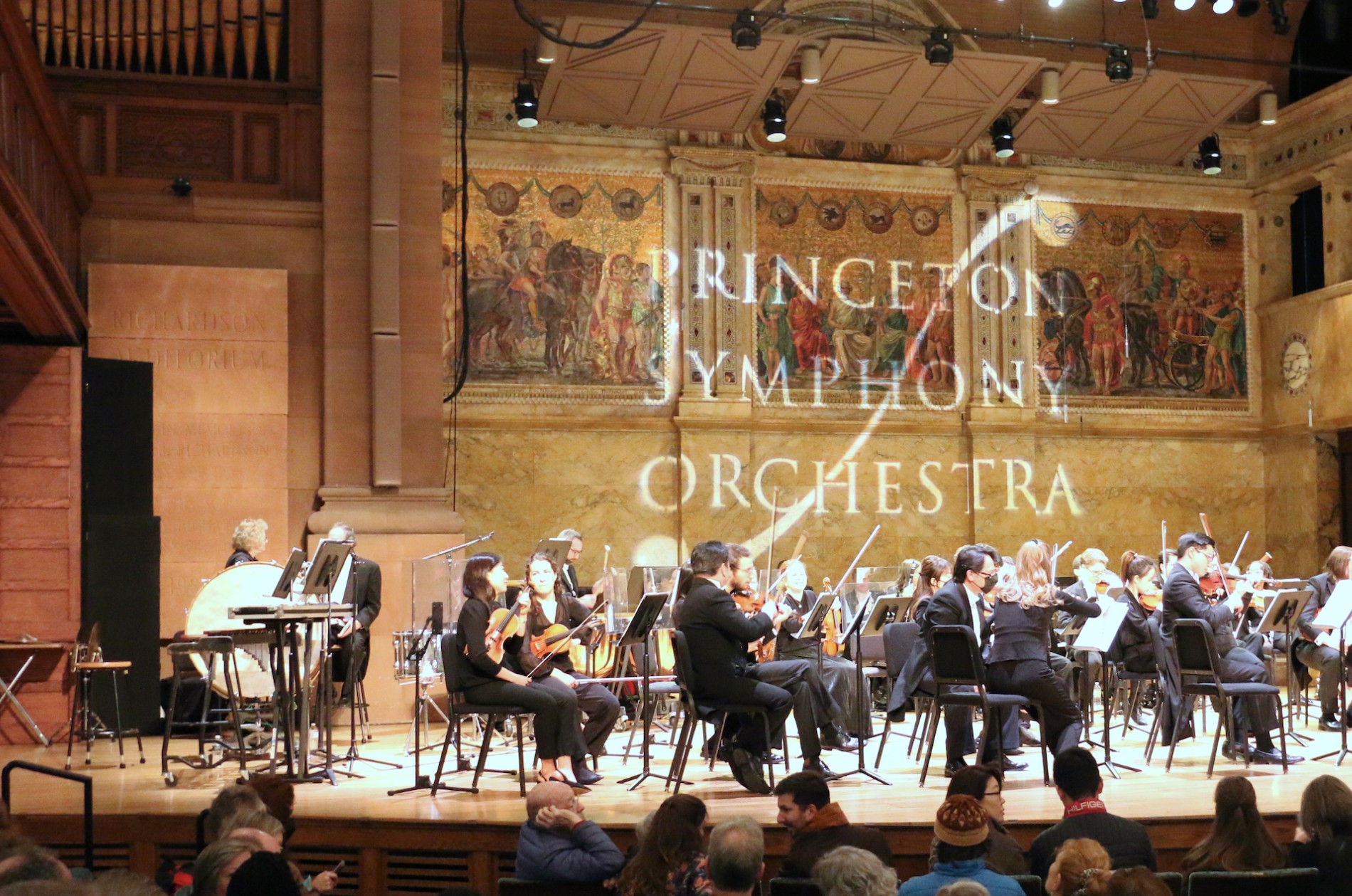 Princeton Symphony Orchestra musicians on stage with PSO logo projected on mosaic wall behind